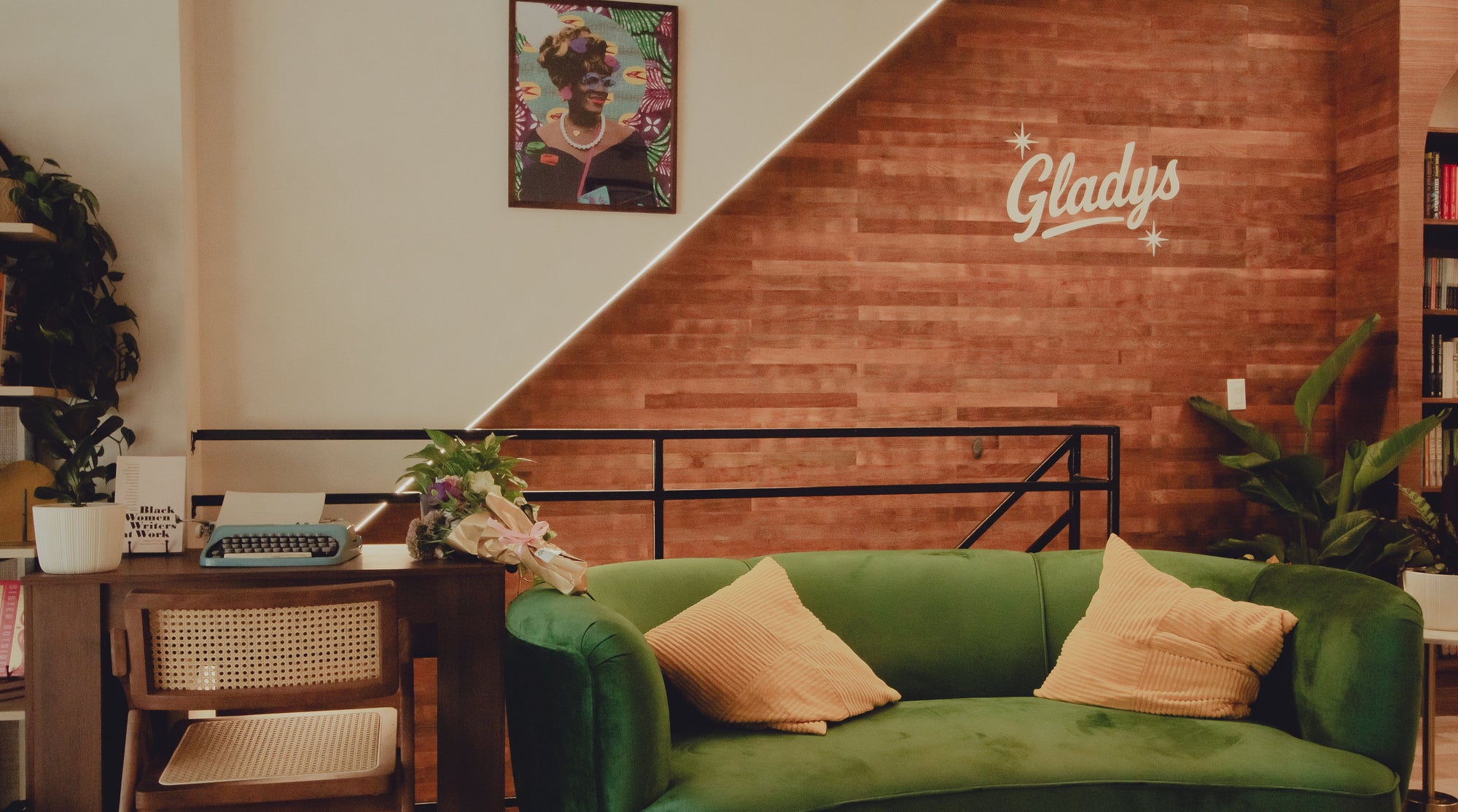 A cozy interior scene featuring a green velvet sofa with two beige pillows in the foreground, a wooden desk with a typewriter and flowers on the left, and a brick accent wall with a white 'Gladys' sign on the right.